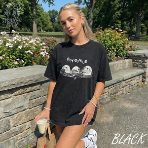 K&ouml;nnte beinhalten: Ein schwarzes Kurzarm-T-Shirt mit einem wei&szlig;en Grafikdruck von drei aus dem Wasser ragenden Robbenk&ouml;pfen. Der Text "BOY G3N1US" ist in Wei&szlig; &uuml;ber der Robbenillustration aufgedruckt. Das T-Shirt hat einen Rundhalsausschnitt und eine lockere Passform.