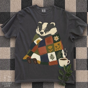 May include: A charcoal gray t-shirt featuring a graphic illustration of a badger wearing round glasses and wrapped in a colorful patchwork quilt. The quilt displays various patterns including a sun, flowers, and leaves. A steaming mug sits nearby.