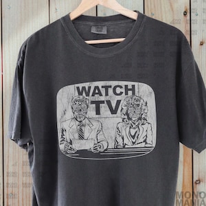May include: Dark grey t-shirt with a graphic that reads "WATCH TV". The graphic shows two figures with skeletal faces, like news anchors. The t-shirt is displayed on a wooden hanger.