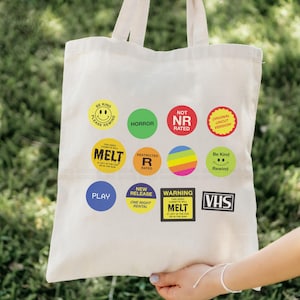 May include: A cream-colored tote bag featuring colorful circular stickers with text like "HORROR," "MELT," and "VHS." The bag is held up by a person. The stickers have various colors and designs, including rainbows and smiley faces.
