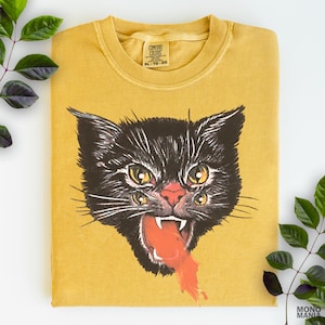 May include: Mustard yellow t-shirt featuring a black cat with four eyes and an open mouth with orange liquid. The shirt has a crew neck and the Comfort Colors brand tag. The graphic is centered on the front.