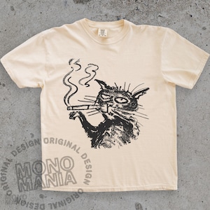 May include: Cream-colored t-shirt featuring a black illustration of a cat smoking a cigarette. The cat has a winking eye and a slightly grumpy expression. The shirt has the words "ORIGINAL DESIGN" and "MONO MANIA" printed on it.
