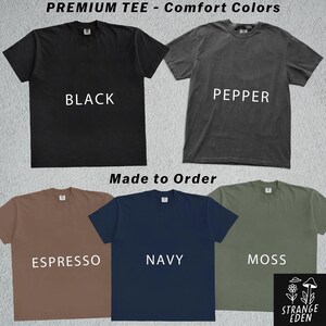 K&ouml;nnte beinhalten: F&uuml;nf kurz&auml;rmelige T-Shirts mit Rundhalsausschnitt in den Farben Schwarz, Pfeffer, Espresso, Marineblau und Moosgr&uuml;n. Der Text PREMIUM TEE - Comfort Colors, Made to Order und der Markenname STRANGE EDEN mit Pilz- und Blumengrafik sind zu sehen.