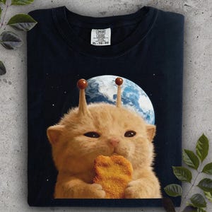 May include: Dark navy t-shirt with a cartoon cat wearing antennae, holding a chicken nugget, against a space and planet background. The shirt has a Comfort Colors tag, size XL-TG-EG.