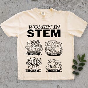 May include: Cream-colored t-shirt with the words "WOMEN IN STEM" in black. Below the text are four black illustrations: spells, tarot, energy, and magic. The shirt has a crew neck and short sleeves.