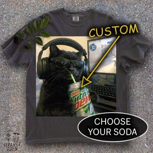 May include: A charcoal gray t-shirt featuring a graphic of a black cat wearing black headphones and sunglasses while drinking from a Diet Mountain Dew can with a straw. The graphic includes yellow arrows pointing to the soda can with the text CUSTOM.