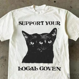 Peut inclure: T-shirt blanc cassé avec un motif noir d'un chat à deux têtes. Le texte "SUPPORT YOUR" est au-dessus de l'image du chat, et "LOCAL GOVEN" est en dessous. Le t-shirt a un col rond et des manches courtes.