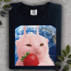 Pode incluir: Camiseta azul marinho com uma criatura rosa parecida com um gato segurando um morango vermelho em frente a um fundo da Terra azul e branco. A camiseta tem uma etiqueta Comfort Colors. A criatura tem duas antenas.