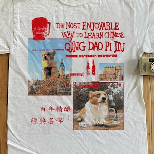 May include: White t-shirt with a graphic design featuring a corgi dog and beer bottles. The text on the shirt reads "THE MOST ENJOYABLE WAY TO LEARN CHINESE: QING DAO PI JIU" in red. Additional Chinese text is also present.