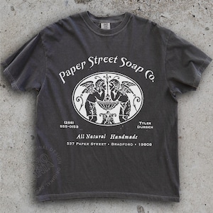 May include: Charcoal grey t-shirt with a white graphic. The graphic includes the text "Paper Street Soap Co." and an oval design with two figures. Additional text includes a phone number, "All Natural Handmade," and an address.