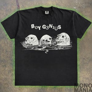 Peut inclure: T-shirt noir avec un motif blanc de trois phoques avec le texte "Boy G3NIUS" imprimé sur le devant.