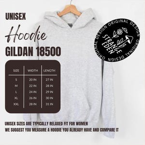 Puede incluir: Una sudadera con capucha gris jaspeado claro con bolsillo tipo canguro. El texto incluye UNISEX Hoodie GILDAN 18500, una tabla de tallas con medidas en pulgadas y un logotipo circular que dice STRANGE EDEN ORIGINAL DESIGN con un gr&aacute;fico de flor y seta.
