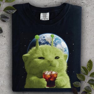 May include: Navy blue t-shirt featuring a green alien cat holding a glass of cola with ice. The cat has antennae and is set against a backdrop of Earth and stars. The shirt has a Comfort Colors tag.