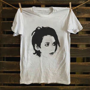 Op de afbeelding: Wit T-shirt met een zwart-wit grafisch portret van een vrouw. Het T-shirt hangt aan een waslijn. De afbeelding is een gecentreerde afbeelding op de voorkant van het T-shirt. Het T-shirt is gemaakt van zacht materiaal.