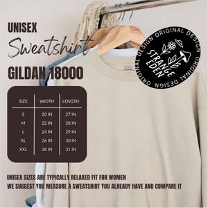 Puede incluir: Una sudadera unisex Gildan 18000 de color beige colgada en una percha de madera. Una tabla marr&oacute;n oscuro muestra las tallas de S a XXL con medidas de ancho y largo en pulgadas. Un logotipo negro circular presenta una flor, un hongo y el texto Strange Eden.
