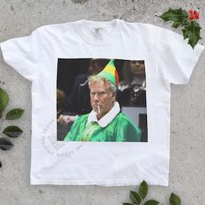May include: White t-shirt featuring a humorous image of a person dressed as an elf, smoking a cigarette. The person is wearing a green jacket, a white collar, and a colorful elf hat. The shirt has the words "ORIGINAL DESIGN" printed on it.