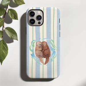 Può includere: Custodia per telefono con sfondo a righe bianche e azzurre. Presenta un'illustrazione ad acquerello di due lontre che si abbracciano, circondate da forme blu e verdi simili all'acqua. Le lontre sono in tonalità marroni e beige.