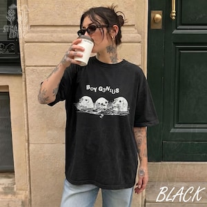K&ouml;nnte beinhalten: Ein schwarzes Kurzarm-T-Shirt mit einem wei&szlig;en Grafikdruck von drei aus dem Wasser ragenden Robbenk&ouml;pfen. &Uuml;ber den Robben steht der Text BOY G3N1US in einer stilisierten Schriftart. Das T-Shirt hat einen Rundhalsausschnitt und eine lockere Passform.