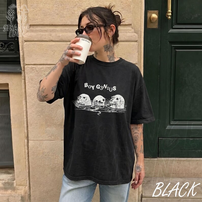K&ouml;nnte beinhalten: Ein schwarzes Kurzarm-T-Shirt mit einem wei&szlig;en Grafikdruck von drei aus dem Wasser ragenden Robbenk&ouml;pfen. &Uuml;ber den Robben steht der Text BOY G3N1US in einer stilisierten Schriftart. Das T-Shirt hat einen Rundhalsausschnitt und eine lockere Passform.