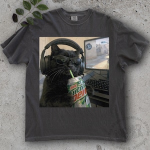 Puede incluir: Camiseta gris oscuro con un gato negro con auriculares y gafas, sosteniendo una lata de Diet Mountain Dew con una pajita. El gato está frente a una pantalla de ordenador. La camiseta tiene un aspecto vintage.