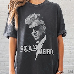 Puede incluir: Camiseta gris oscuro con un retrato en blanco y negro de un hombre con gafas de sol. El texto "STAY WEIRD." está impreso en una fuente gótica debajo. La camiseta tiene mangas cortas y un corte holgado.