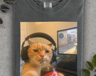 May include: A gray Comfort Colors t-shirt featuring a meme of an orange cat wearing headphones and drinking from a Coca-Cola bottle. The cat is in front of a computer monitor. The Coca-Cola logo is red and white.