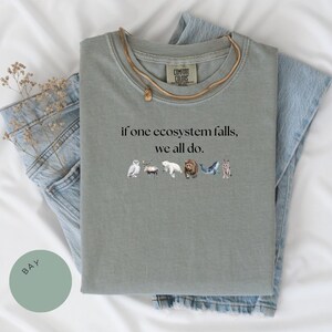 May include: A sage green t-shirt with the text "if one ecosystem falls, we all do." printed on the front. The text is surrounded by small illustrations of animals, including an owl, a deer, a polar bear, a bear, a whale, and a wolf.