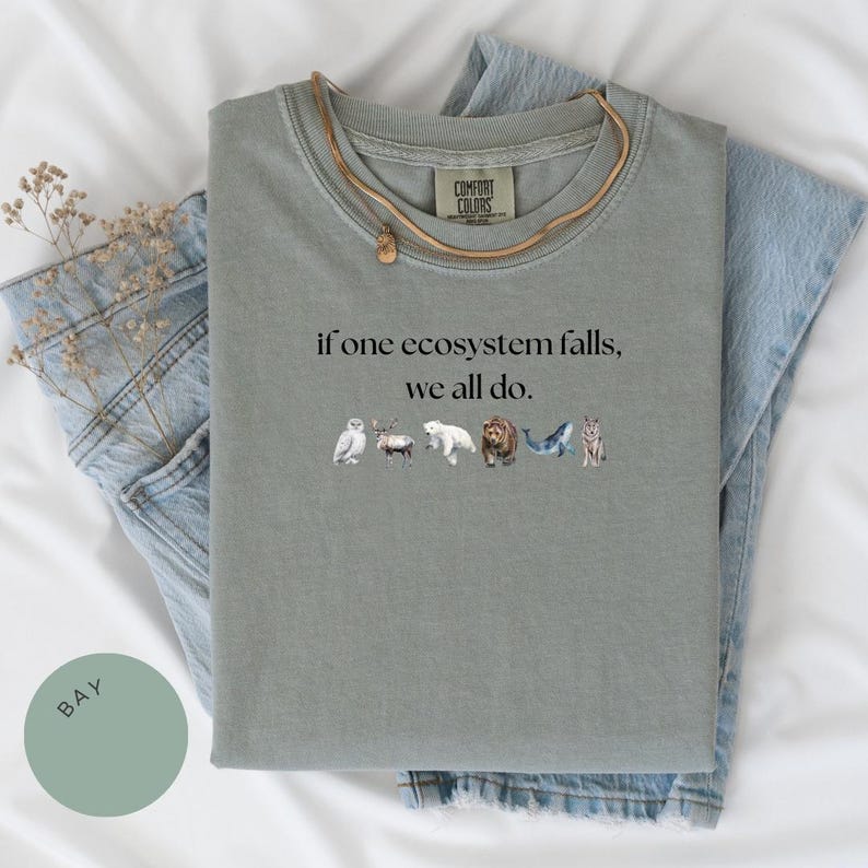 May include: A sage green t-shirt with the text "if one ecosystem falls, we all do." printed on the front. The text is surrounded by small illustrations of animals, including an owl, a deer, a polar bear, a bear, a whale, and a wolf.