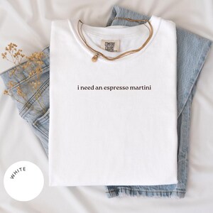May include: White t-shirt with the phrase "i need an espresso martini" printed in black. The shirt is folded and placed on top of a pair of blue jeans. A gold necklace is draped over the shirt.