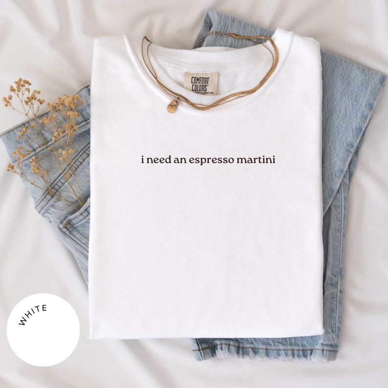May include: White t-shirt with the phrase "i need an espresso martini" printed in black. The shirt is folded and placed on top of a pair of blue jeans. A gold necklace is draped over the shirt.