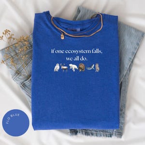 May include: A royal blue t-shirt with the text "if one ecosystem falls, we all do."  The text is in white and is centered on the shirt.  There are small images of animals below the text, including an owl, a reindeer, a polar bear, a bear, a whale, and a wolf.