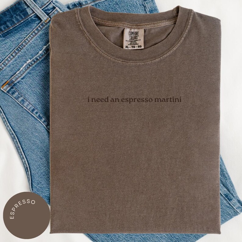 May include: A brown t-shirt with the text "i need an espresso martini" in a simple font. The shirt is folded on top of blue denim jeans. The shirt has a tag that says "Comfort Colors" and the size is XL-TG-EG.
