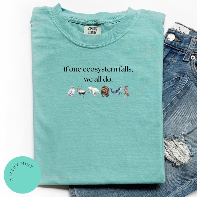May include: A teal blue t-shirt with the text "if one ecosystem falls, we all do." printed on the front. The text is in black and is surrounded by a row of small animal illustrations.