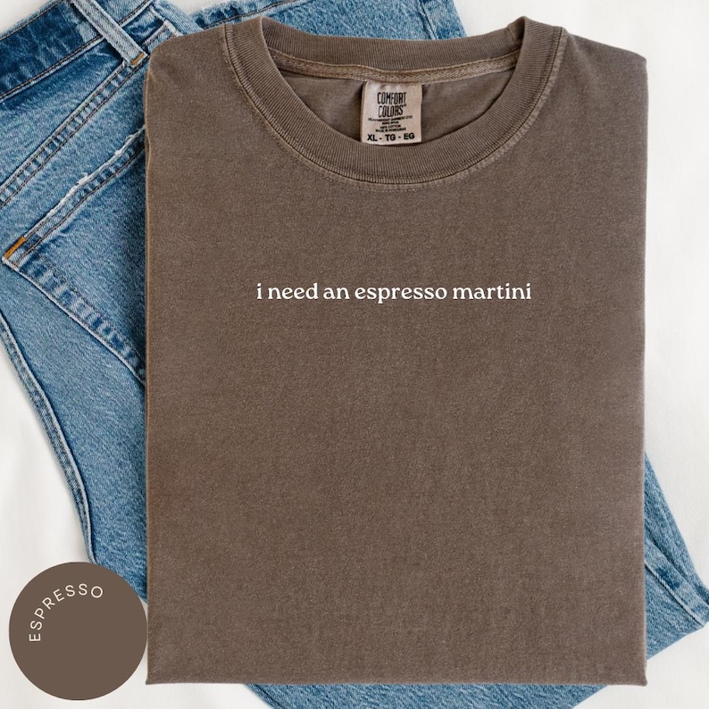 May include: A brown t-shirt with the text "i need an espresso martini" in white. The shirt is folded on top of blue denim jeans. The shirt has a Comfort Colors tag in the upper center.
