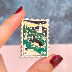 May include: A rectangular enamel pin in the shape of a postage stamp, featuring a black and white orca whale swimming in green water. The pin has a lighthouse, windmill, and the text "9¢".