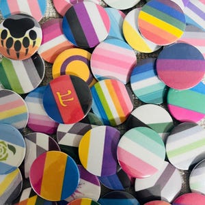 May include: A collection of circular pin-back buttons featuring various colorful pride flag patterns, including horizontal stripes, vertical stripes, and a paw print design. One button displays a yellow Greek pi symbol on a red, blue, and black background.