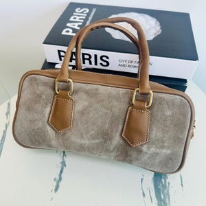 May include: A rectangular handbag with a suede-like gray body and brown leather trim. The bag features a top handle and two decorative leather pieces with gold-colored hardware. The word "PARIS" is visible on a book in the background.