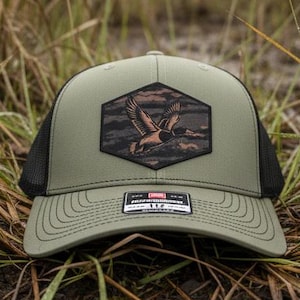 May include: Olive green trucker hat with a black mesh back. The front features a hexagonal patch with a brown and black graphic of a flying duck. The hat is resting on the ground amidst tall grass.