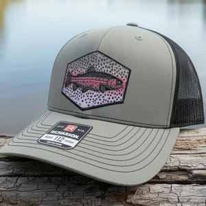 May include: A gray and black trucker hat with a fish graphic. The hat features a pink and red fish design inside a hexagonal patch. The hat is sitting on a wooden log near a body of water.