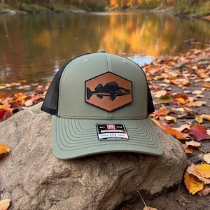 May include: A green and black trucker hat with a fish patch. The hat features a mesh back and a curved brim. The patch is a brown hexagon with a black fish design. The hat is sitting on a rock.