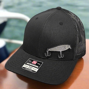 May include: Black trucker hat with a mesh back and a curved brim. The hat features a silver fishing lure emblem and a Richardson 112 Trucker patch. The hat is adjustable.