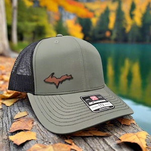 May include: Olive green trucker hat with a black mesh back and a brown patch of the Upper Peninsula of Michigan. The hat has a curved brim and a Richardson 112 tag. The hat is sitting on a wooden surface with fall leaves.