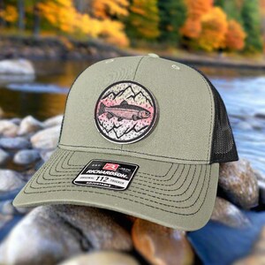 May include: A green and black trucker hat with a patch featuring a trout and mountains. The patch has a pink, white, and blue color scheme. The hat has a Richardson 112 label.