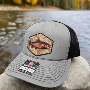 May include: A gray and black trucker hat with a camouflage patch featuring a trout. The patch is a hexagon shape with a brown trout on a tan and brown camouflage background. The hat has a black mesh back and a gray front with a black stitched brim. The hat has a black label with white text that reads "EST. 1970 RICHARDSON. ORIGINAL 112 TRUCKER ADJUSTABLE".