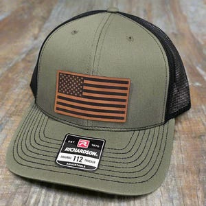 May include: A green and black trucker hat with a brown leather American flag patch. The hat has a black mesh back and a black stitched brim. The hat has a label that reads "EST. 1970 RICHARDSON ORIGINAL 112 TRUCKER ADJUSTABLE".