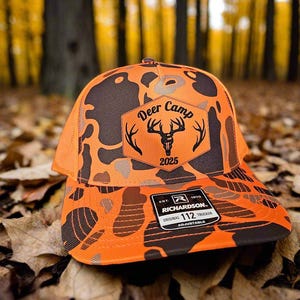 May include: An orange and brown camouflage trucker hat with the text "Deer Camp 2025" and a deer head logo. The hat has a mesh back and a curved brim. The tag reads "Richardson Original 112 Trucker Adjustable".