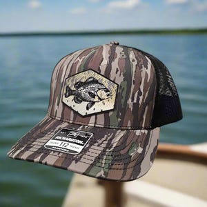 May include: A camouflage trucker hat with a black mesh back. The hat features a black and white embroidered image of a fish on the front. The hat has a black label with the text "Richardson 112 Trucker Adjustable" on the side.