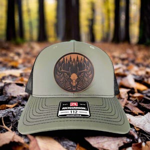 May include: A green and black trucker hat with a brown leather patch featuring a deer skull and forest scene. The patch has the text "Richardson" and "112 Original Trucker" on it.