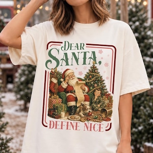 Könnte beinhalten: Ein elfenbeinfarbenes Kurzarm-T-Shirt mit einem Grafikdruck von Santa Claus, der neben einem Weihnachtsbaum und einem Hund eine Liste liest. Der Text Dear Santa, Define Nice steht in roter und grüner Schrift in einem rechteckigen Rahmen.