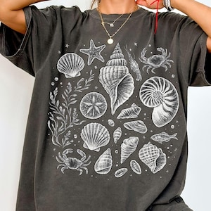 Könnte beinhalten: Dunkelgraues T-Shirt mit einer weißen Illustration verschiedener Muscheln, Seesterne und Meereslebewesen. Das Design umfasst eine große Muschel, eine Krabbe und andere Meeresbewohner. Das Shirt hat eine lockere Passform.